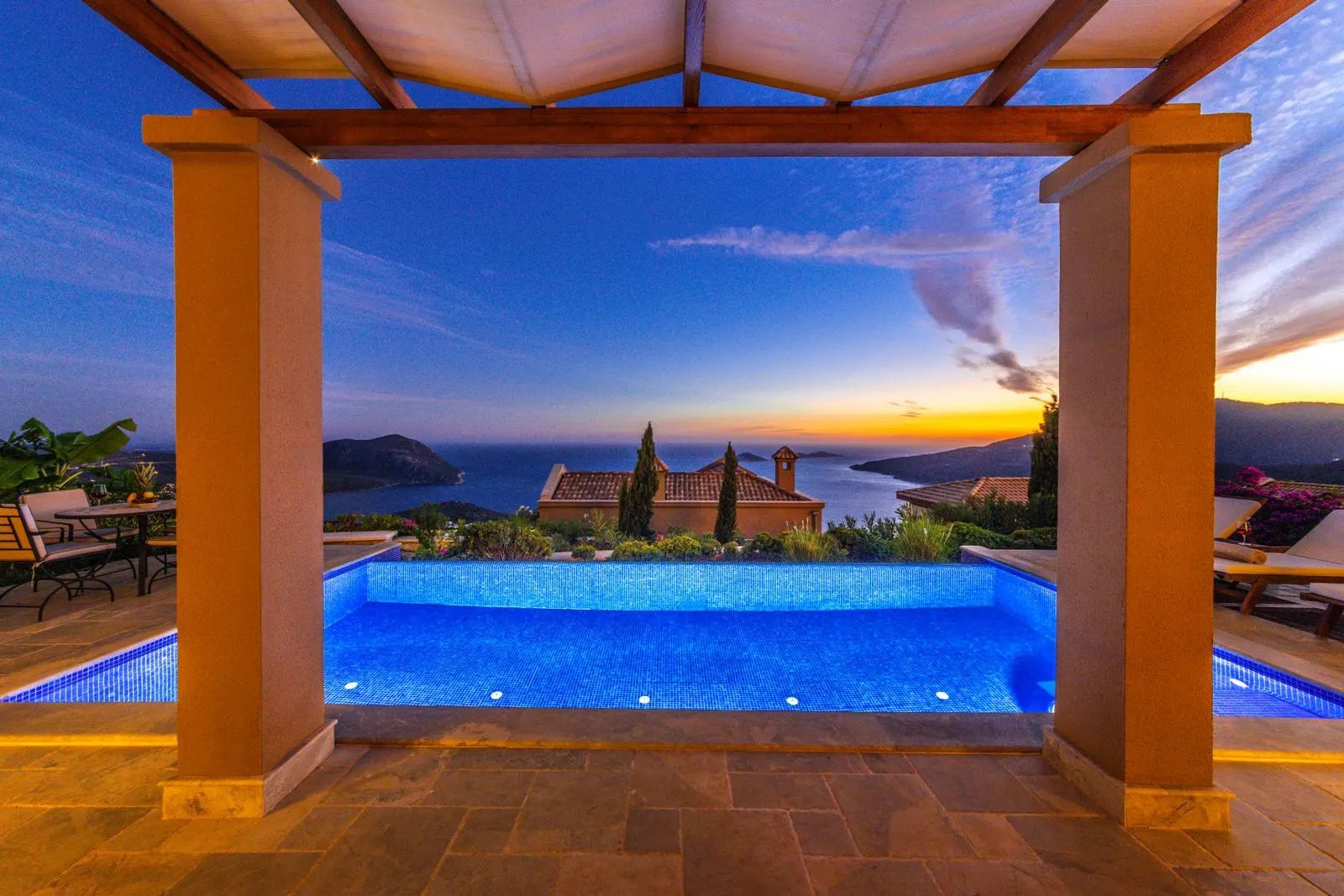 Illuminated infinity pool at dusk, seen from a covered porch, with the sea and coastline beyond