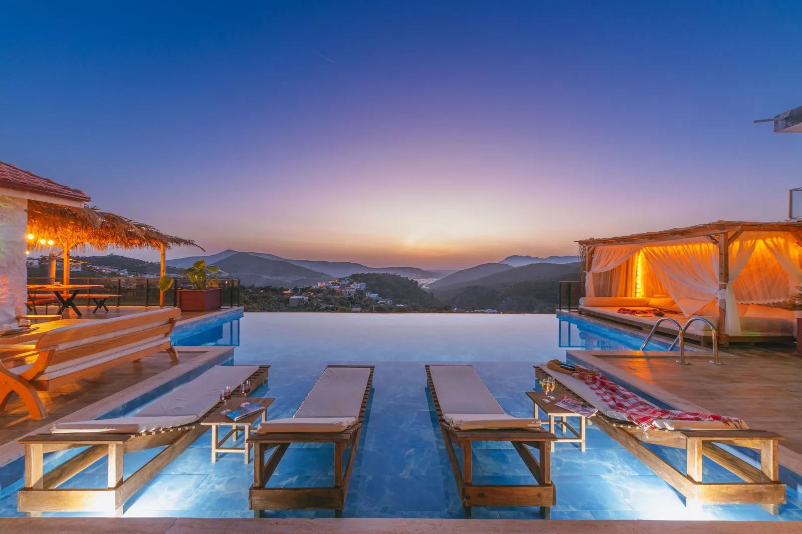 Infinity pool with sun loungers, a pergola and an illuminated canopy bed at sunset, with mountains behind