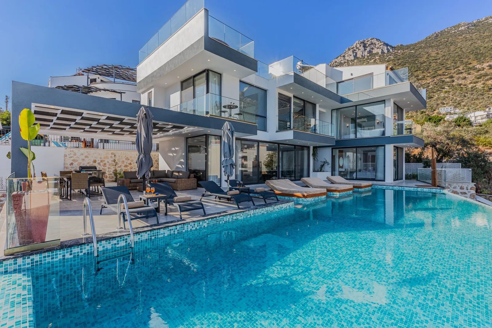 Modern multi-storey villa with glass terraces, sun loungers and a large blue mosaic swimming pool, with a mountain behind